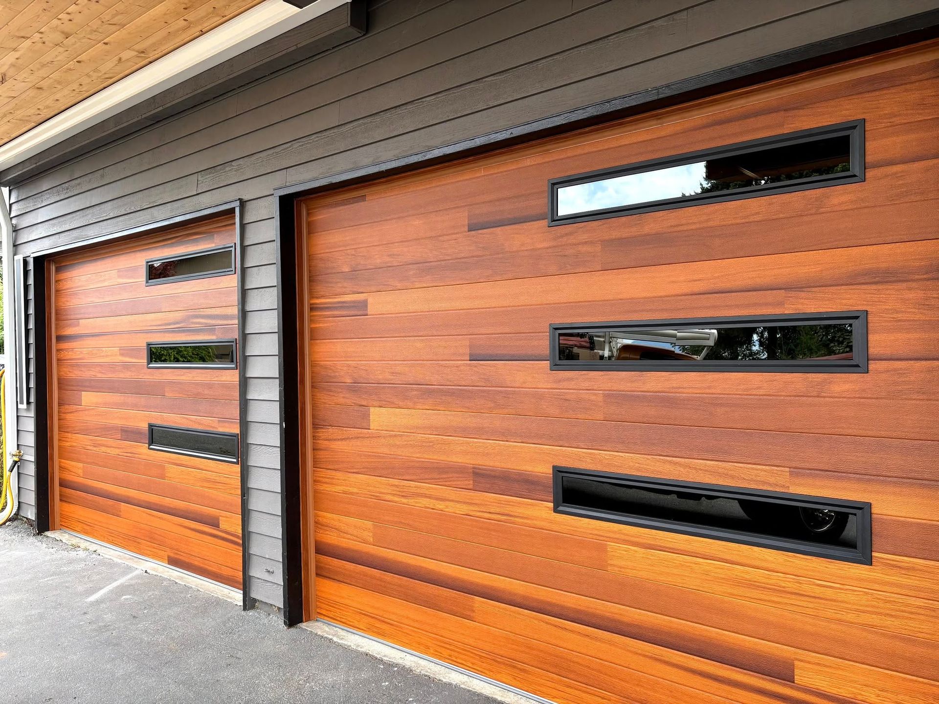 Two modern wooden garage doors with black horizontal windows on a gray driveway.