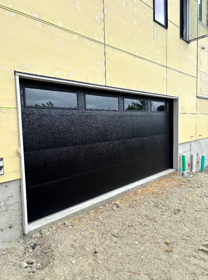 Black garage door on a beige building exterior, viewed from an angle on a gravel lot.