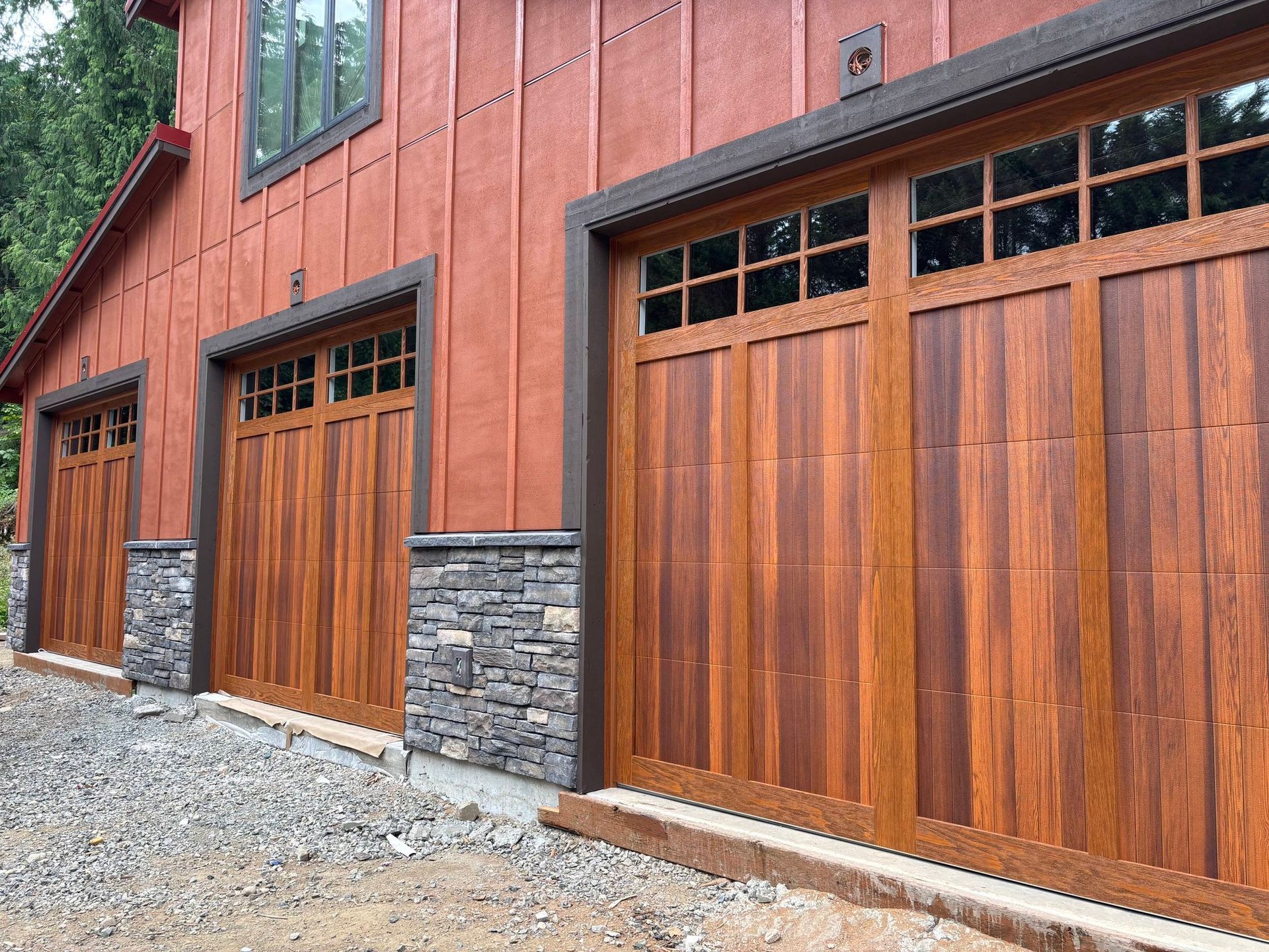 Rustic red building with three wooden garage doors and stone accents on a gravel driveway
