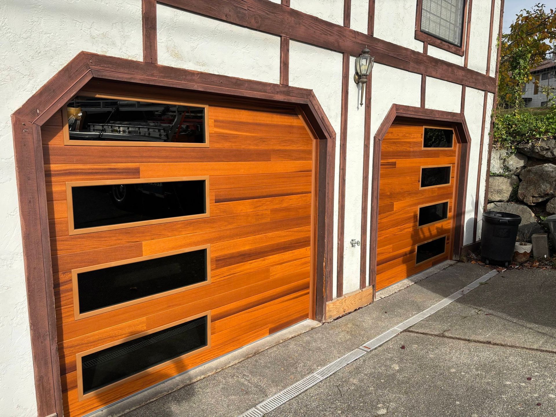 Two wooden garage doors with black windows on a white-and-brown building exterior