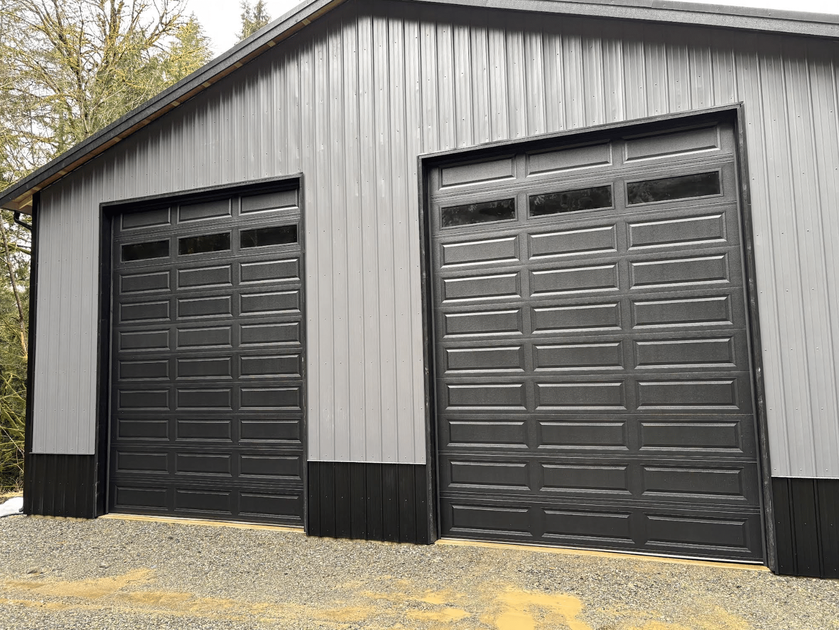 Gray metal garage building with two black roll-up doors and black trim on gravel.