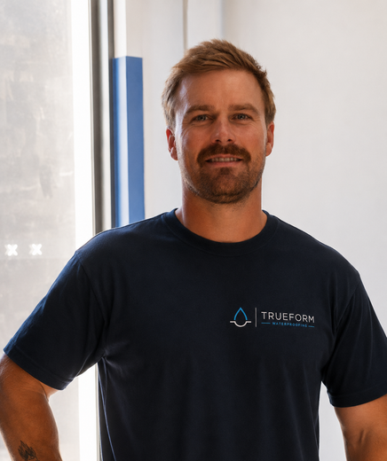 Man in a navy T-shirt standing indoors, smiling in front of a bright window.