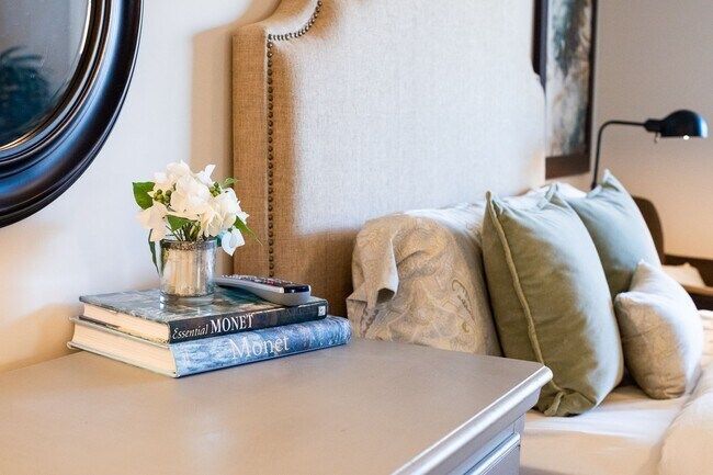 Cozy bedroom corner with a bed, layered pillows, bedside lamp, and flowers on stacked books