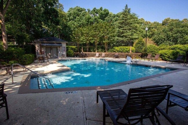 Outdoor swimming pool surrounded by trees, with lounge chairs and a small pool house on a sunny day