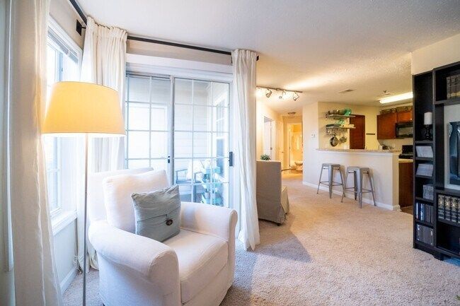 Bright living room with white armchair, floor lamp, sliding glass door, and bar stools in the background