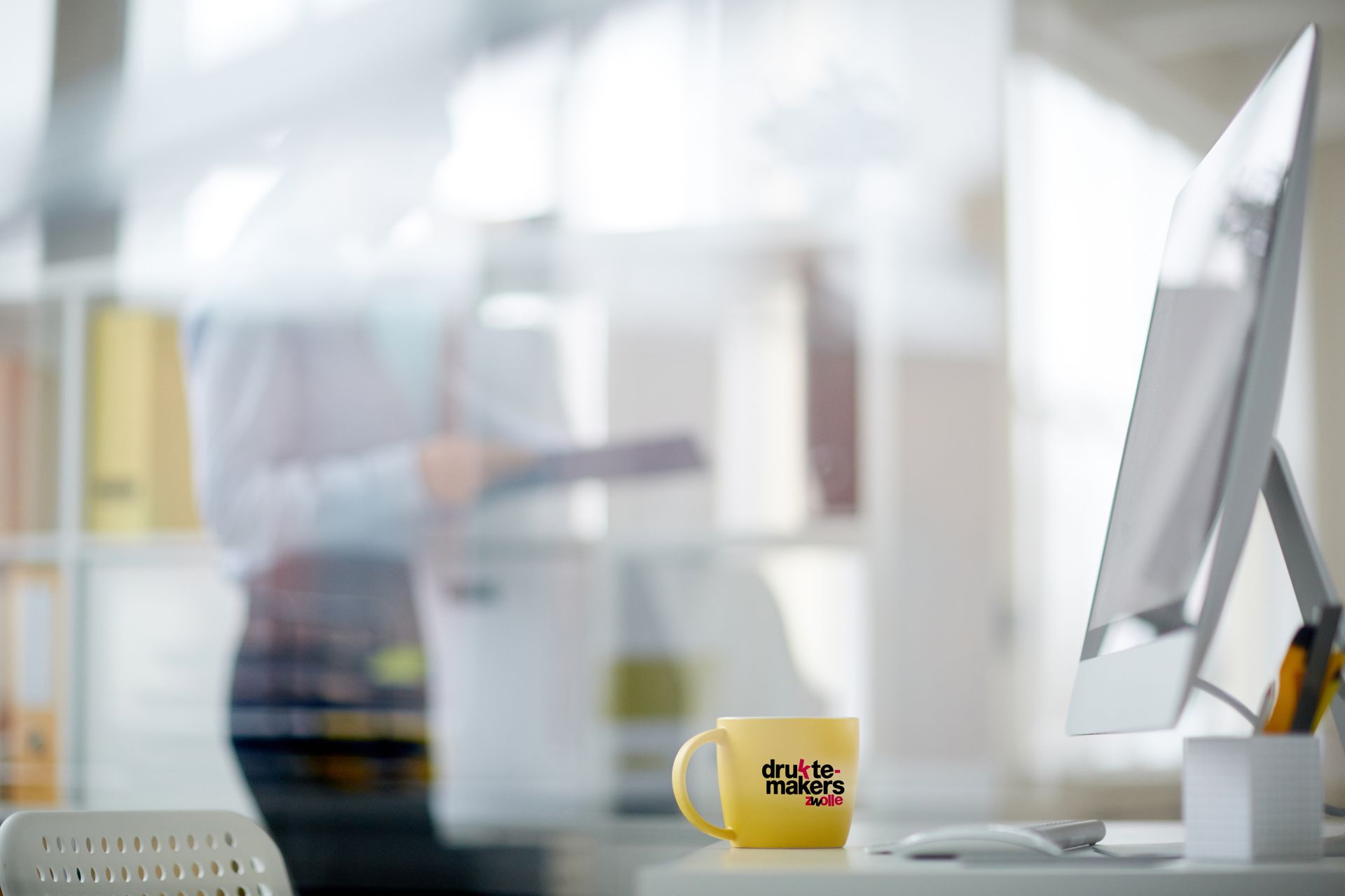 Modern bureau met computermonitor, gele mok en een wazig persoon op de achtergrond.