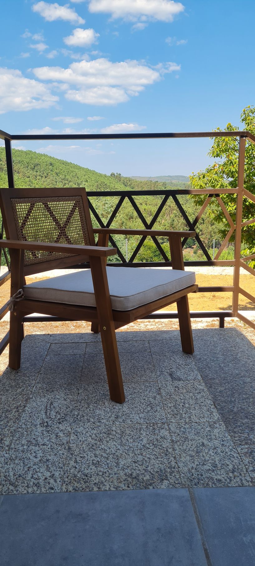 Uma cadeira de jardim em madeira com uma almofada bege, com vista para uma montanha com floresta verdejante.