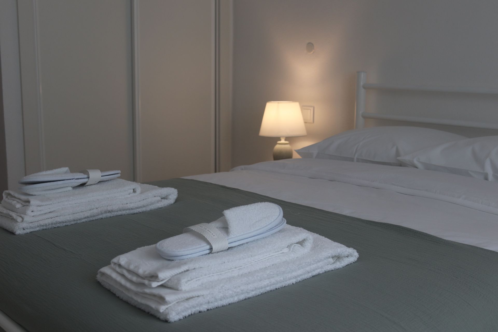 Um quarto com uma cama de casal e um roupeiro. Sobre a cama estão almofadas, toalhas e chinelos. O candeeiro está aceso.