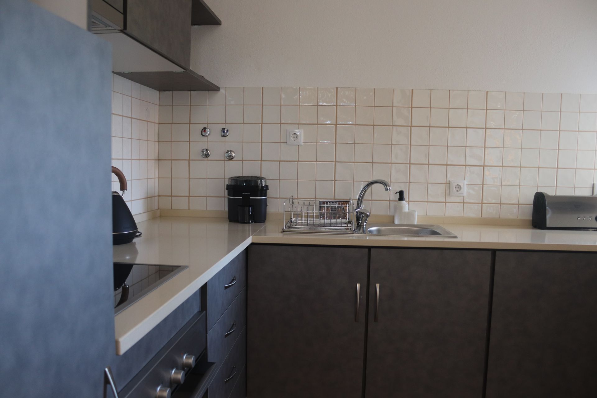 Cozinha com placa elétrica, chaleira, espremedor elétrico, lava loiças e armários. Em tons de branco e cinzento.