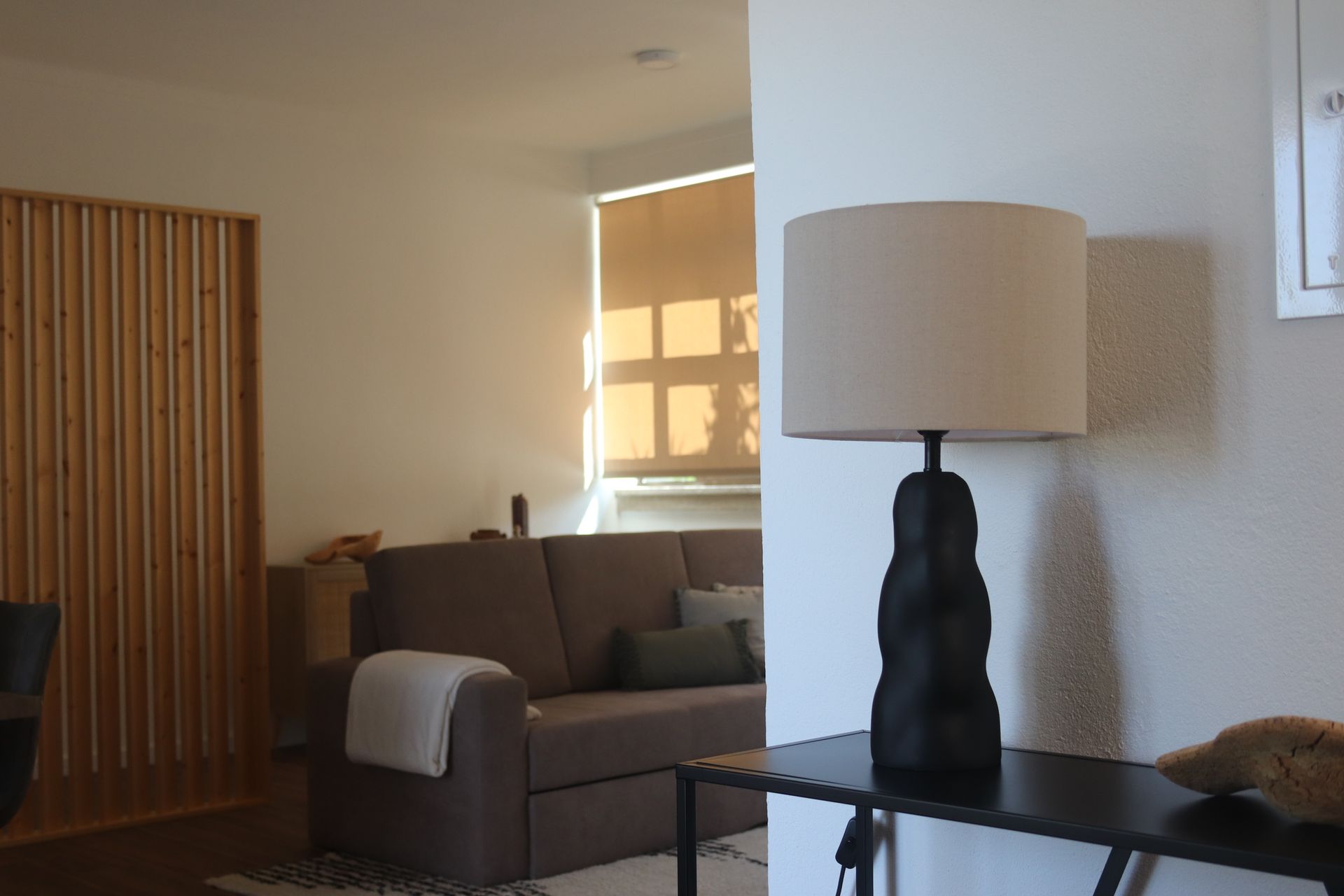 Candeeiro moderno em preto e bege, sobre uma mesa preta na entrada de uma casa, com vista para a sala de estar.