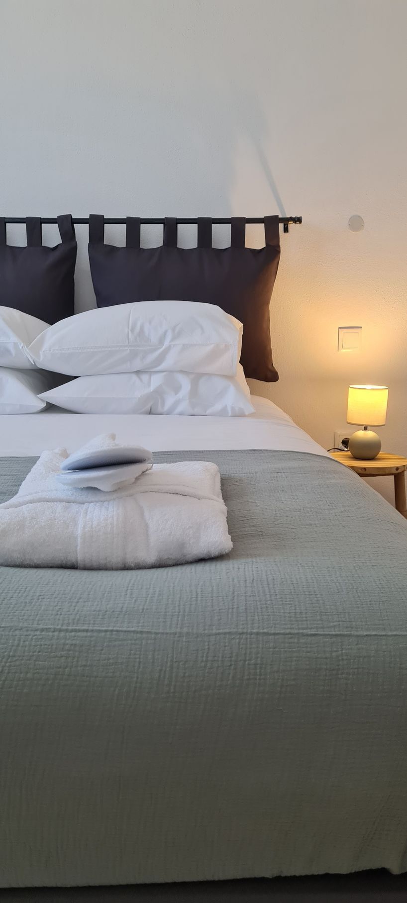 Um quarto com uma cama de casal, com almofadas, robe e chinelos e sobre a mesa de cabeceira tem um candeeiro aceso.