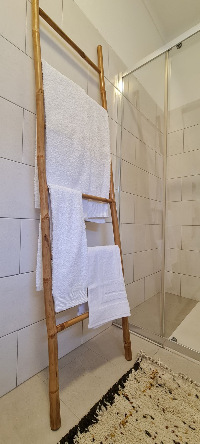 Casa de banho com um toalheiro em bambu, com toalhas de banho brancas de diferentes tamanhos e um tapete confortável no chão.