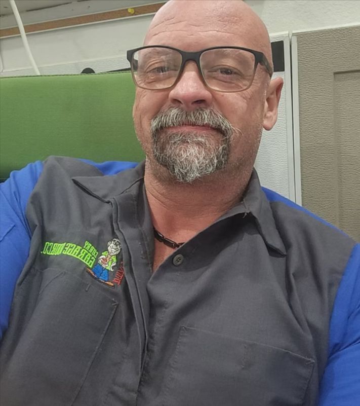 Smiling bald man with glasses wearing a gray-and-blue collared shirt, seated indoors.