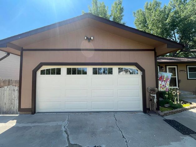Front view of a tan house with a white two-car garage door and cracked driveway under a sunny blue sky