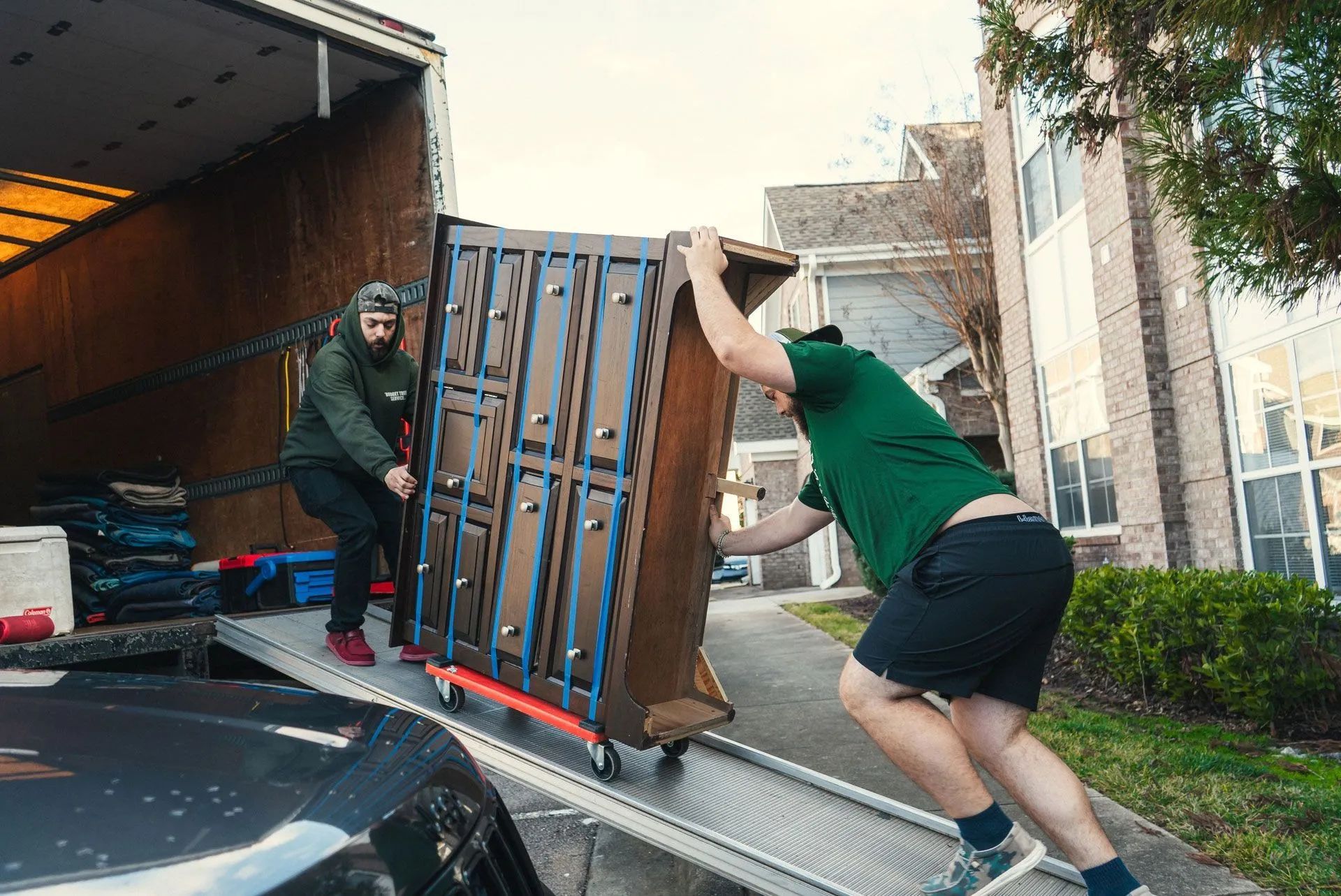 Two workers loading a large wooden cabinet onto a truck ramp outdoors