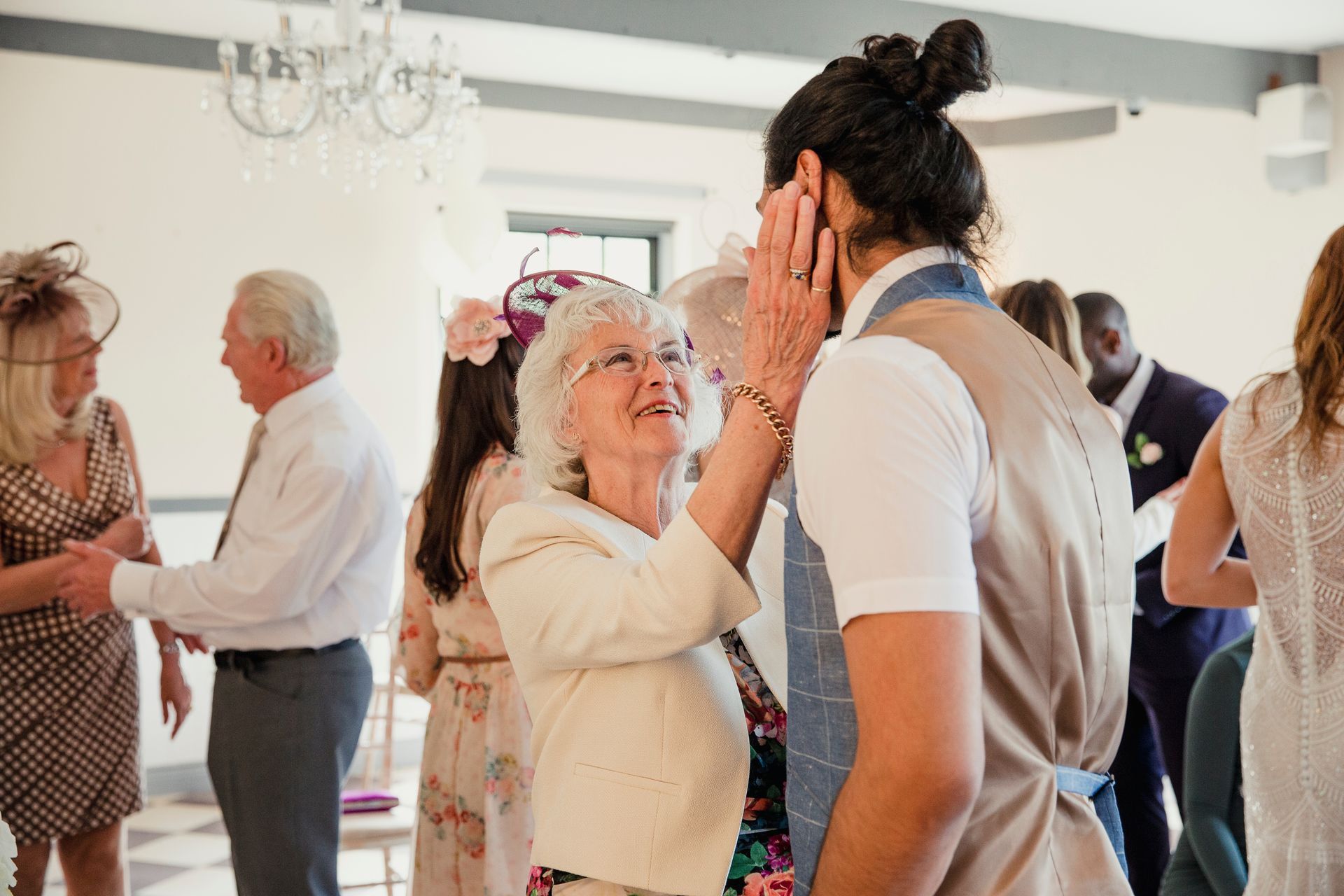 People dancing at a bright indoor social gathering, with an elderly woman smiling and touching a man’s face.