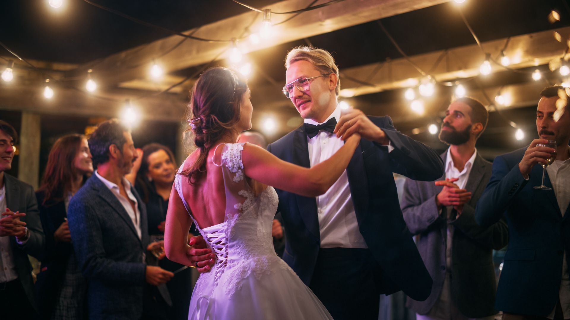 Couple dancing at a wedding reception under warm string lights, surrounded by cheering guests