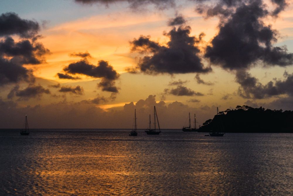 Sunset over calm sea with sailboats, glowing clouds, and a dark island silhouette