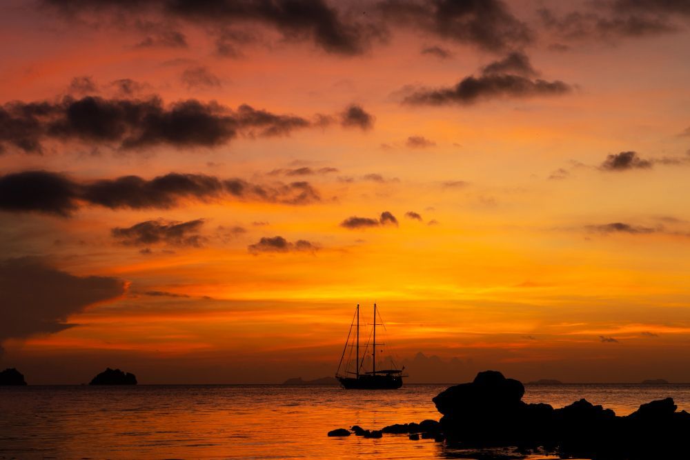Silhouetted sailboat at sunset over calm sea, with dark rocks in the foreground and orange sky.