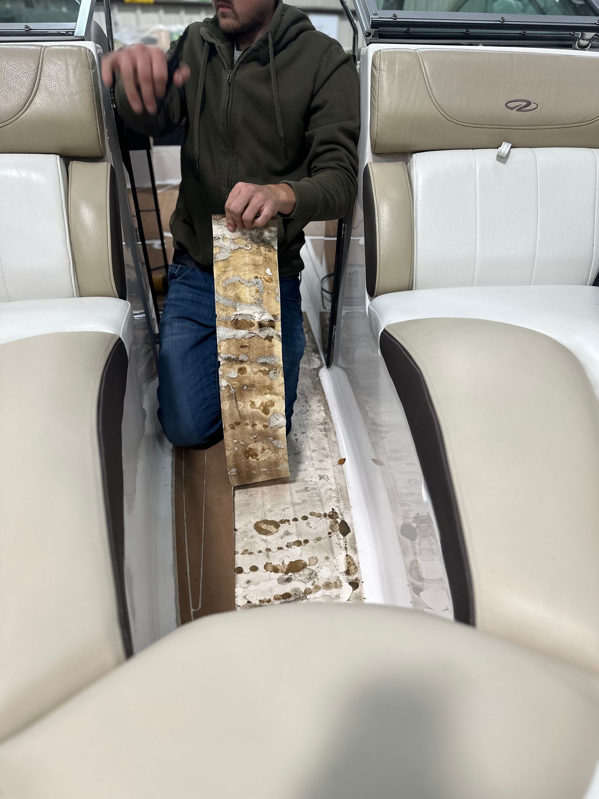 Man kneels in a boat, holding a section of damaged wood flooring. Beige seats line sides.