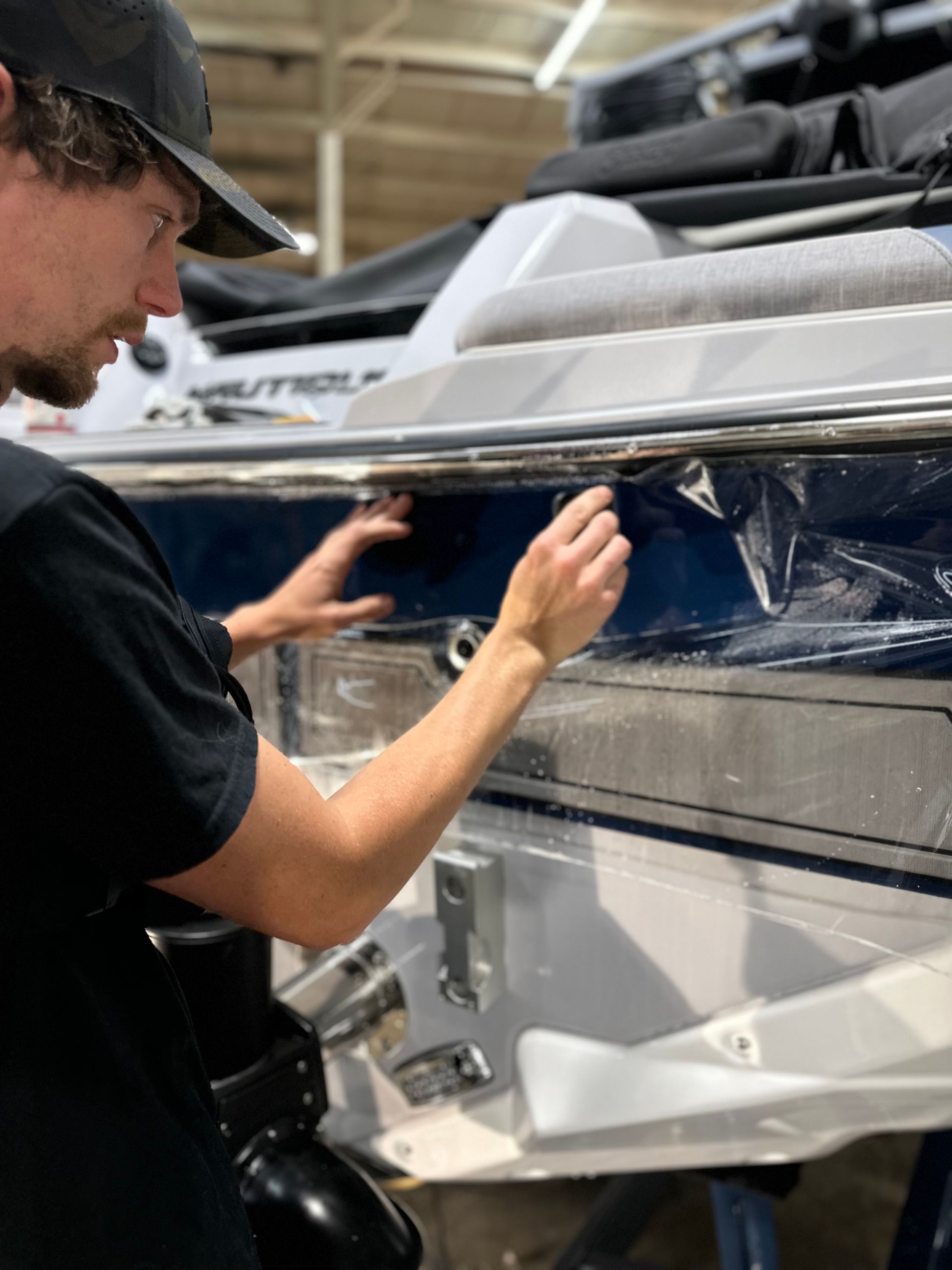 Man applying protective film to a blue boat's side. He's wearing a cap and focused. Inside setting.