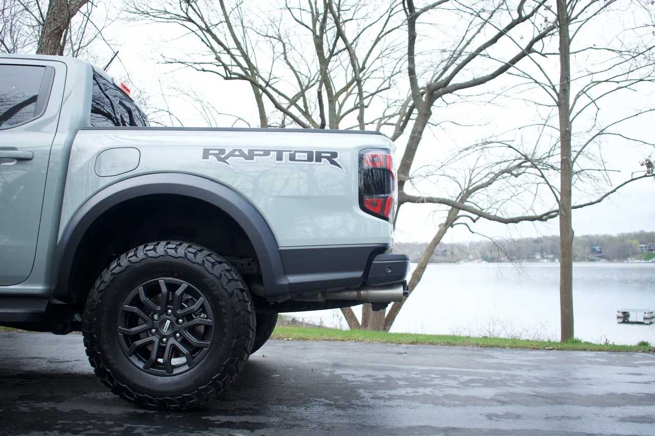 Silver Ford Raptor truck parked lakeside, under cloudy sky, near bare trees.