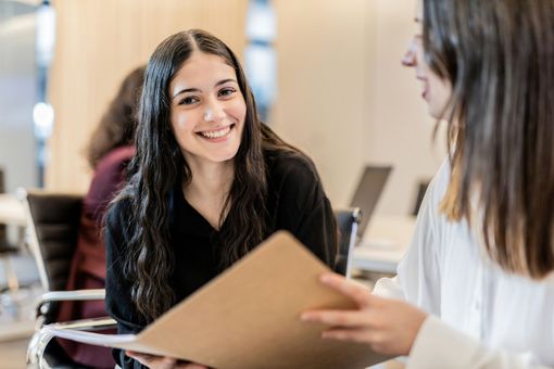 A woman is smiling while holding a folder and talking to another woman.
