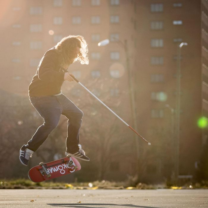 Dan Mancina is captured midair doing a flip trick on his skateboard, cane in hand.