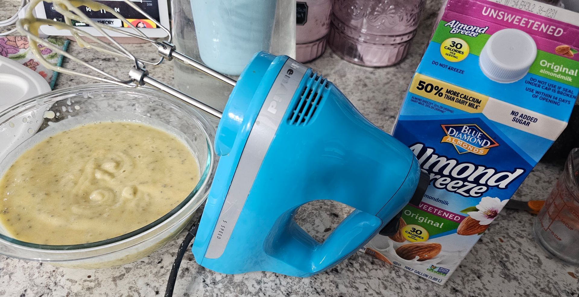 A blue hand mixer sits on a counter next to a bowl of batter and a carton of almond milk.
