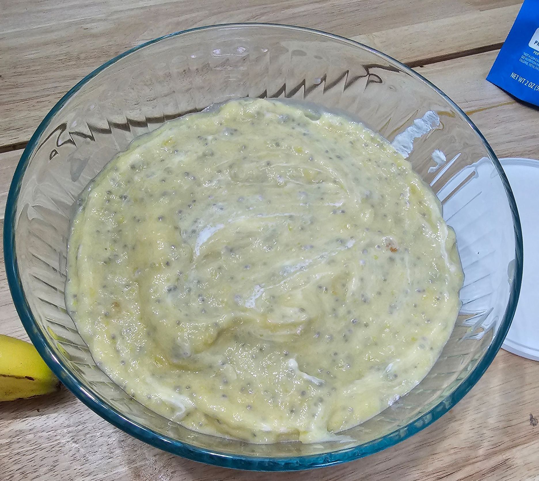 Mashed banana in a glass bowl, next to a banana and a blue packet on a wooden surface.