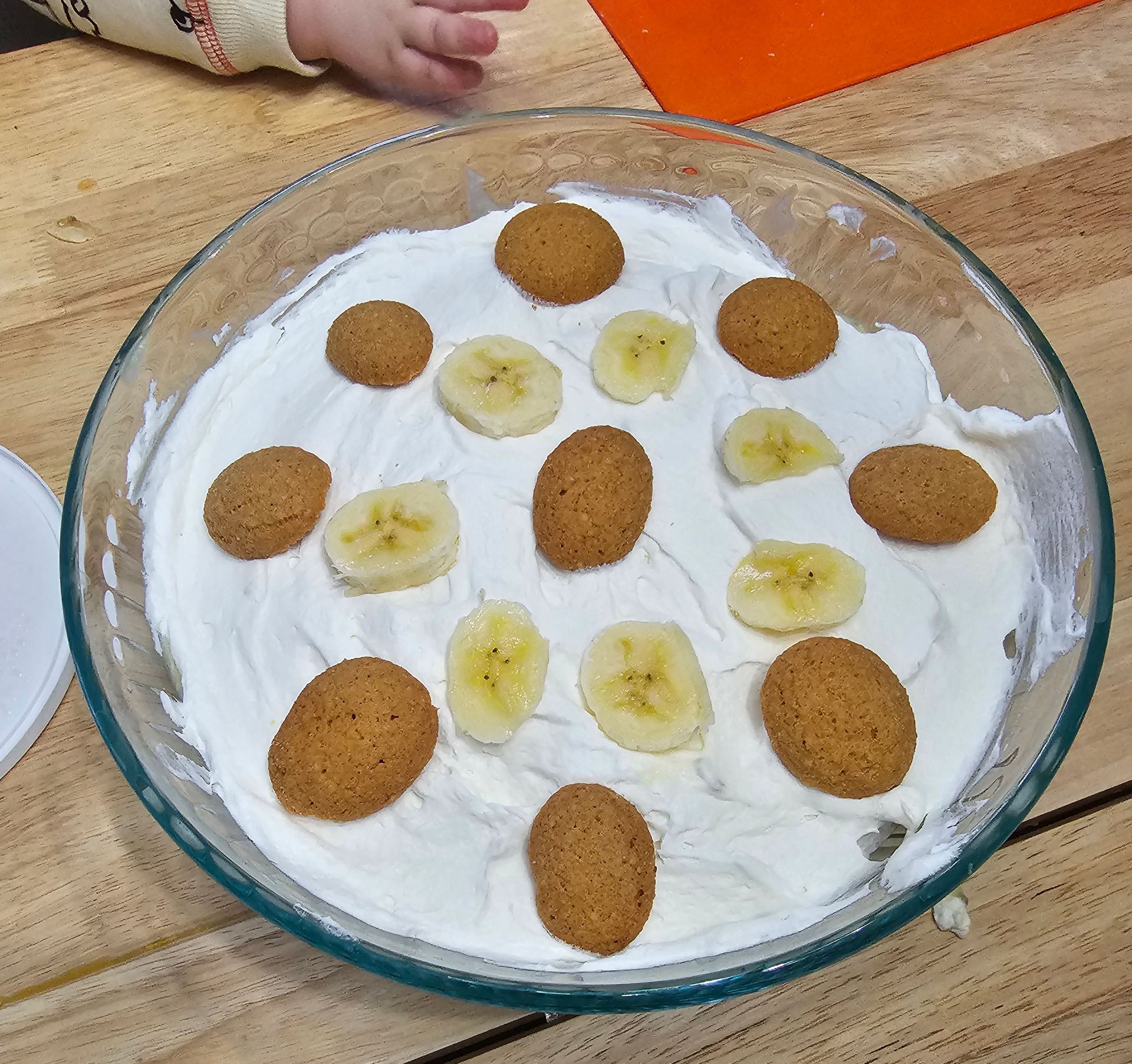 Glass dish with white cream, banana slices, and cookies; small child's hand visible.