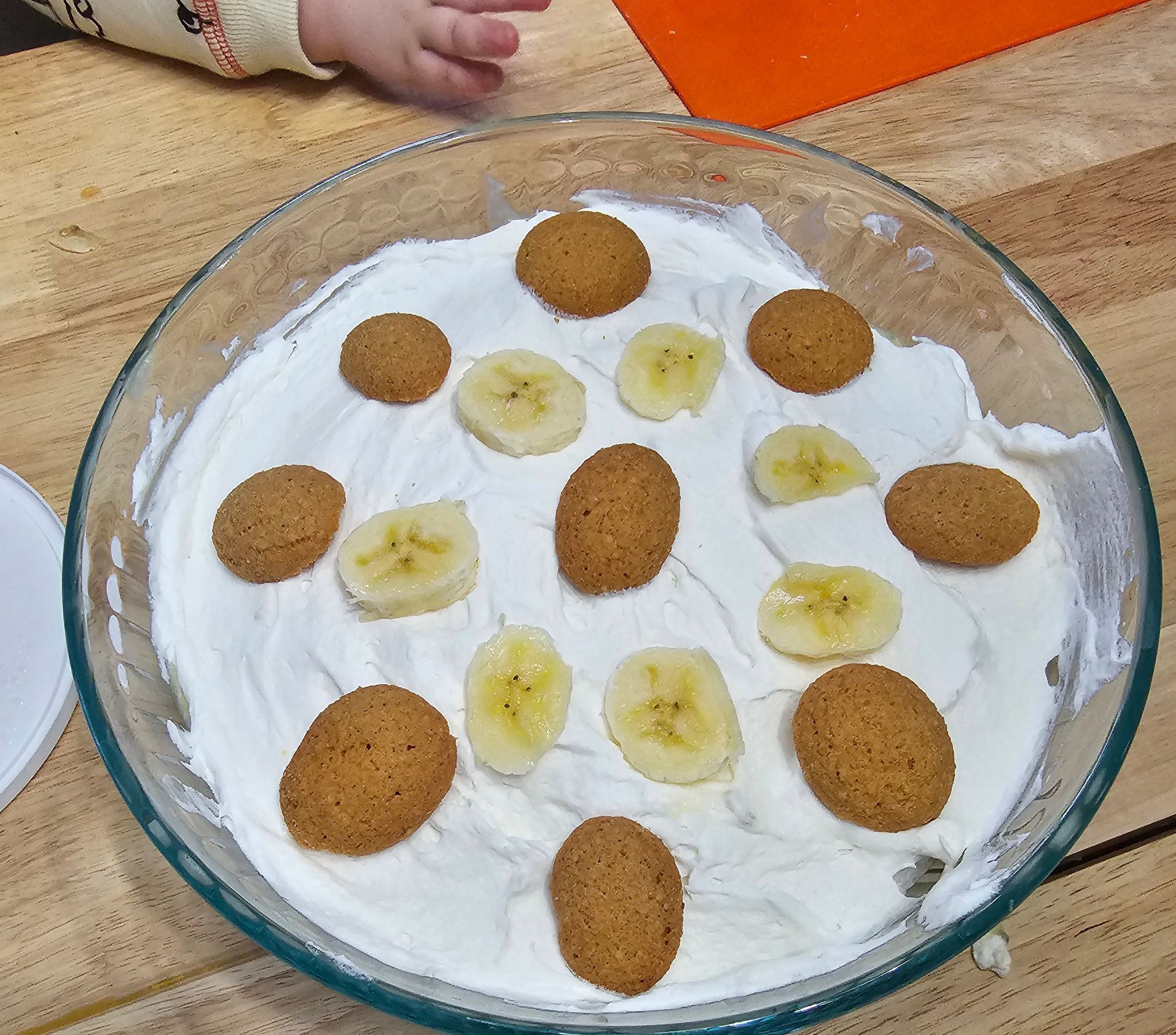 A glass bowl with whipped cream, banana slices, and cookies on a wooden surface.