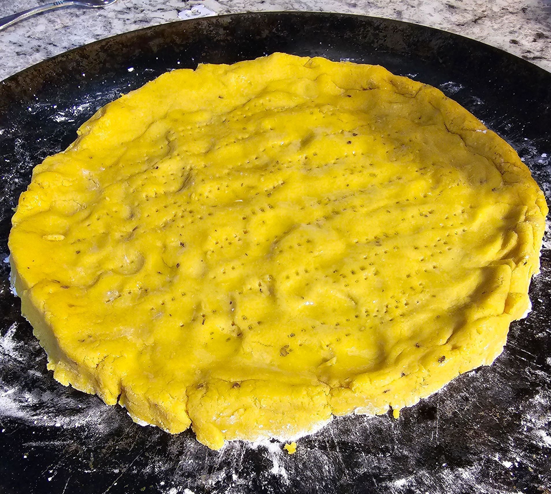Yellow dough circle with small holes on a black pan dusted with flour.