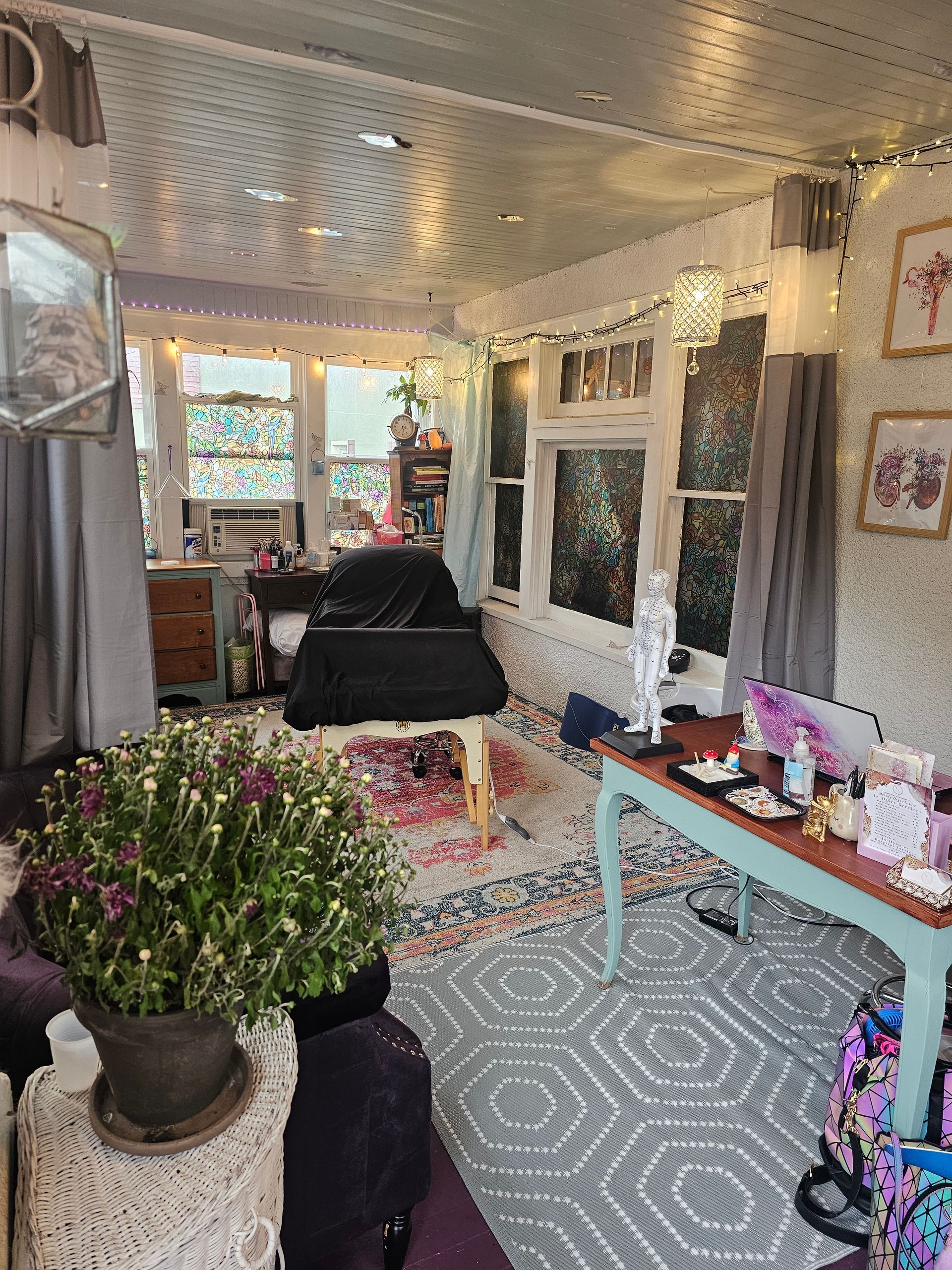 A cozy, eclectic room with a desk, table, and plants. Features a floral floor and string lights.