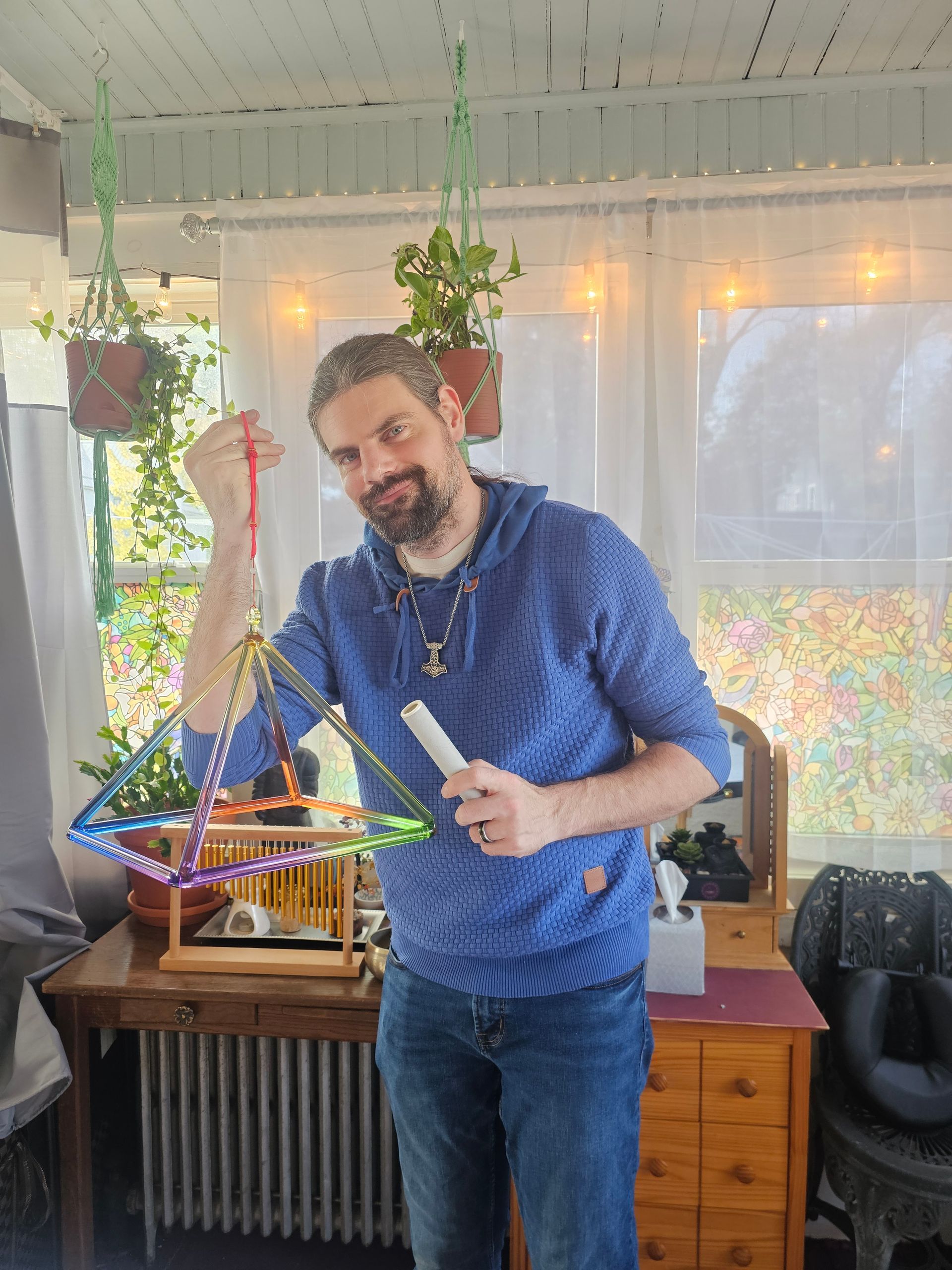 Man with a pyramid-shaped sound device, smiling. Indoors, plants, colorful device, blue hoodie, wooden table, window.