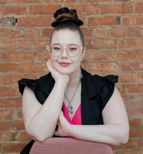 Person with glasses and black vest resting chin on hand, seated against a brick wall.