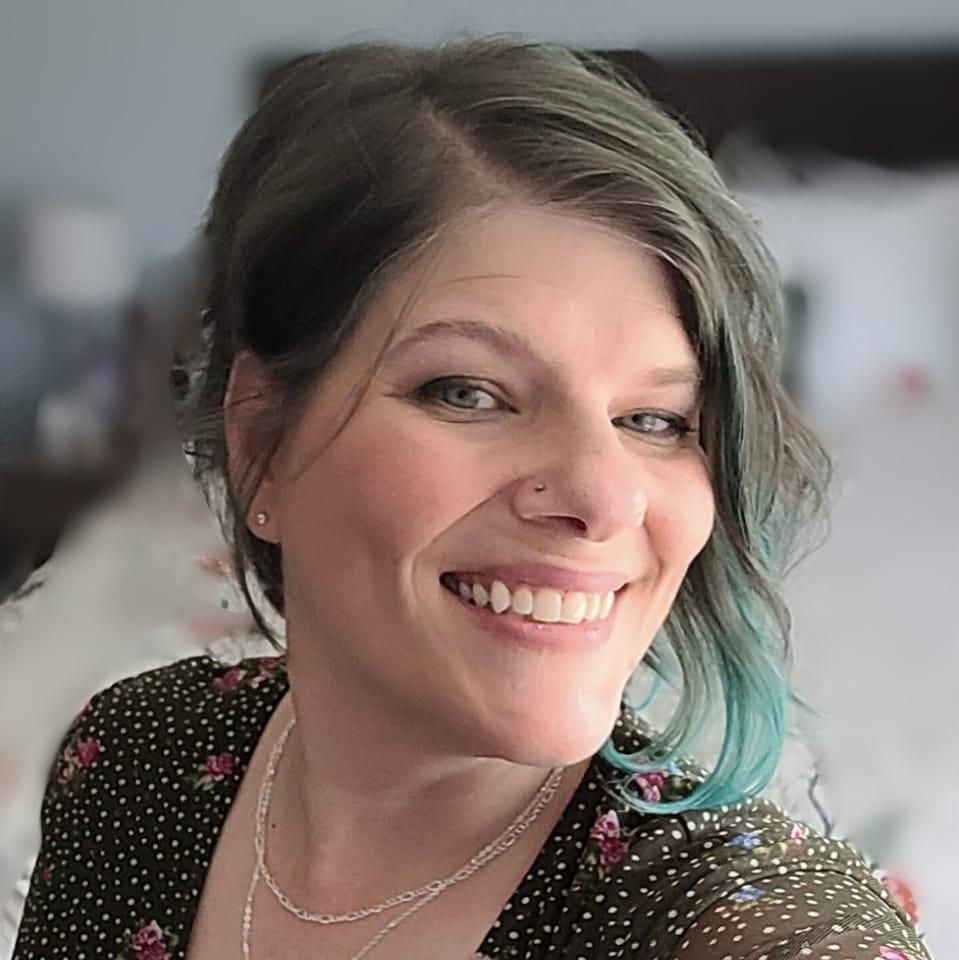 Woman with gray-blue hair smiles, nose ring, wearing a black floral top. Blurred background.