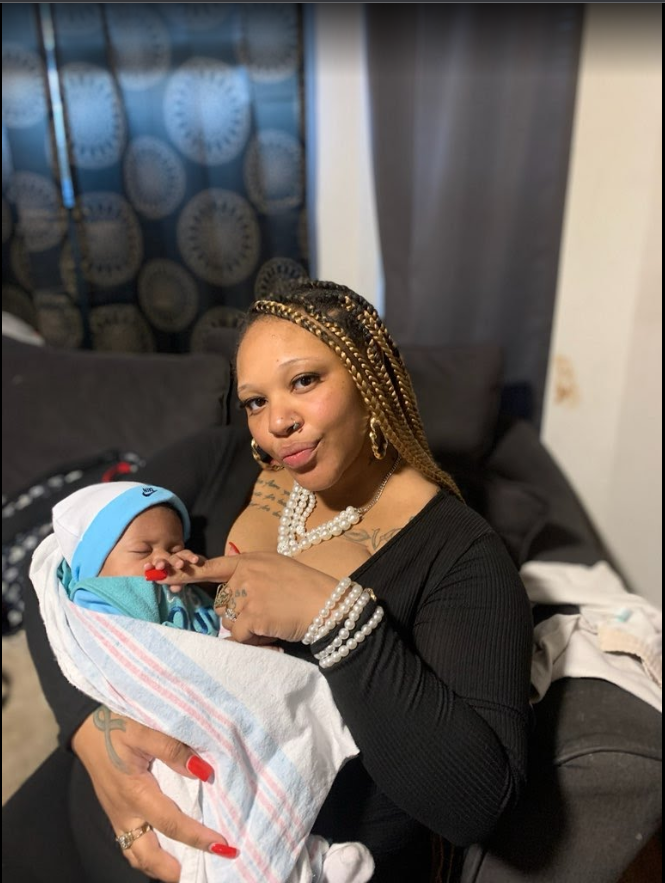 Woman with blonde braids holding a newborn wrapped in a blanket, pointing at its face, on a couch.