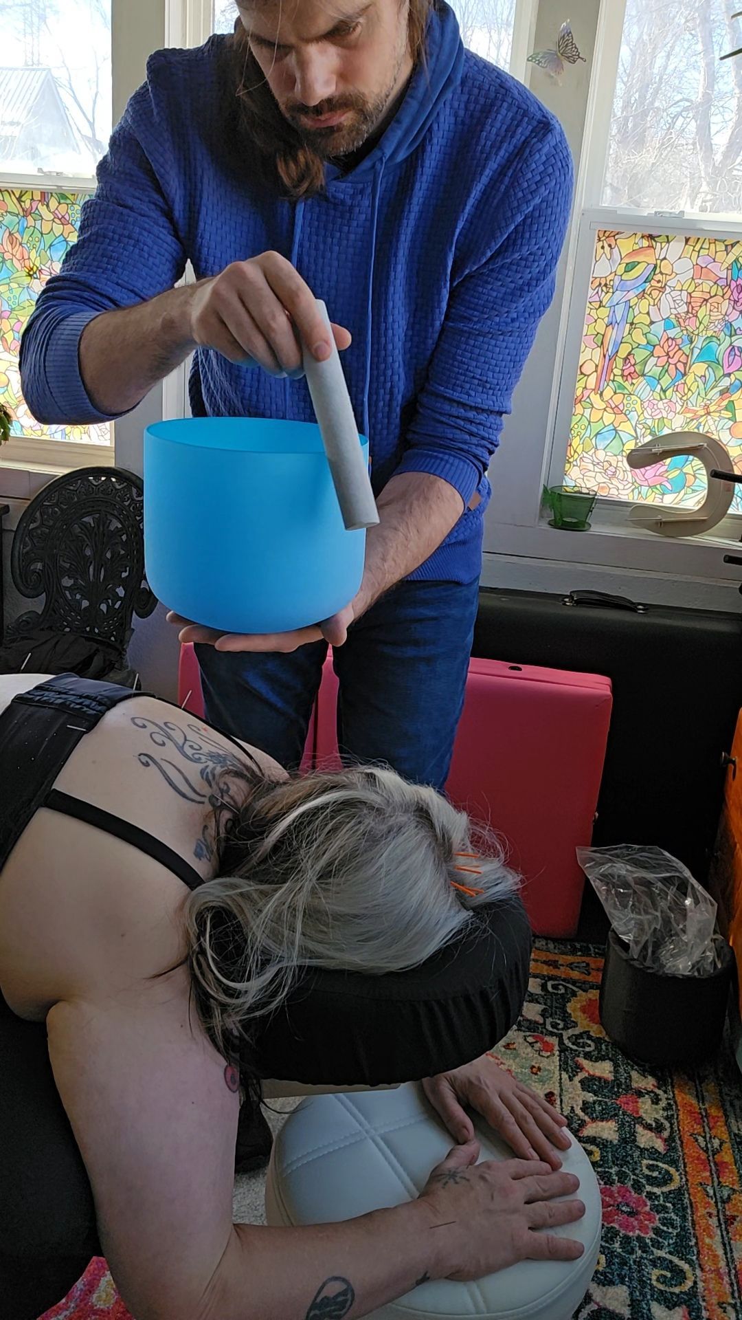 Person holds a mallet to a blue crystal bowl, near another person's head on massage chair.