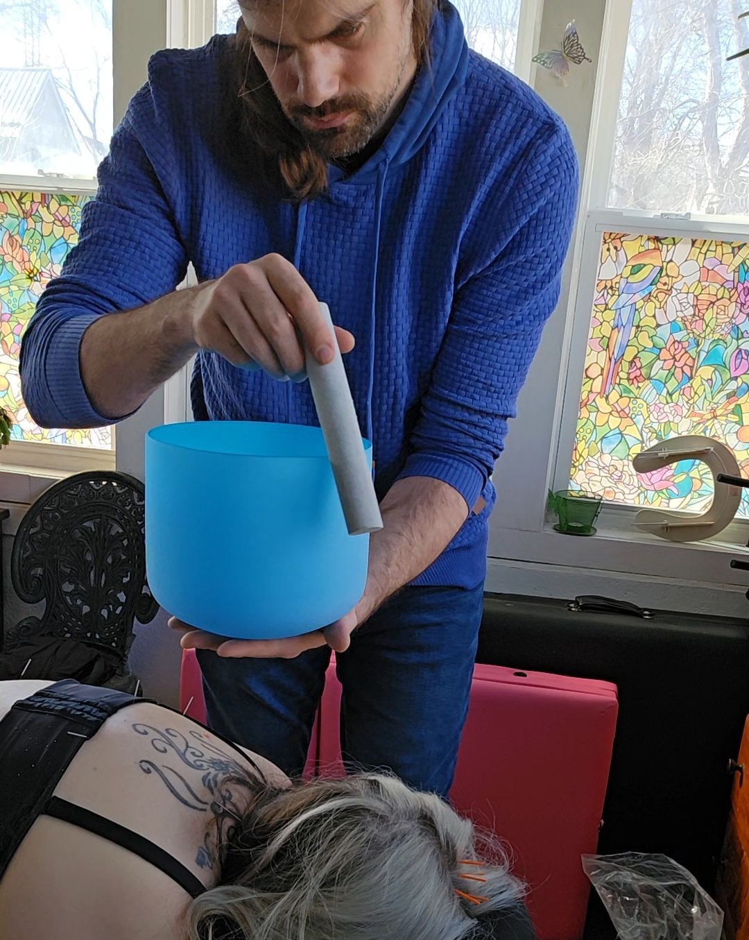 Man plays a blue singing bowl over a person lying down. Indoor setting, colorful window.
