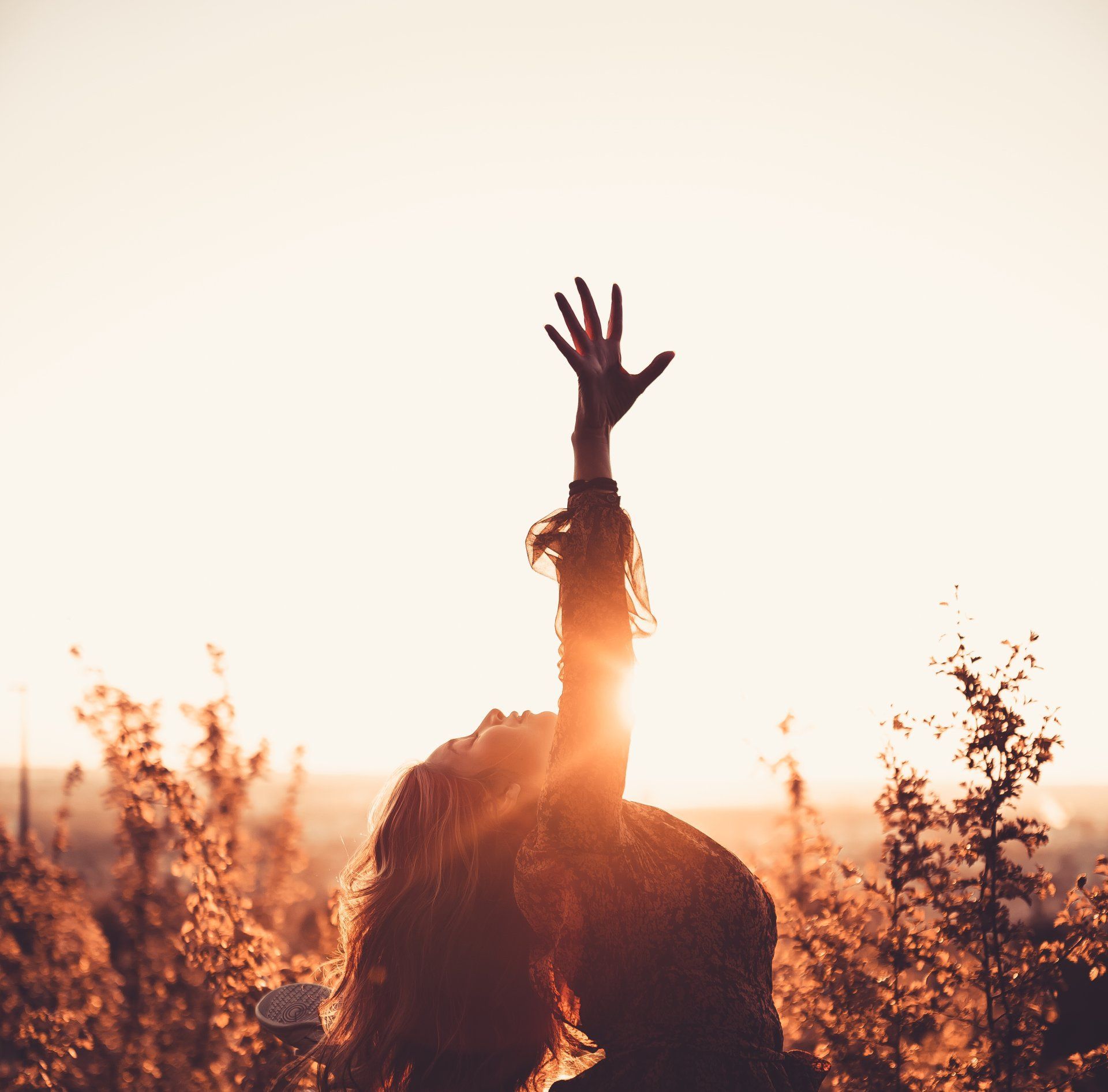 Woman reaches up, arm outstretched towards setting sun in a field of tall grass. Golden light.