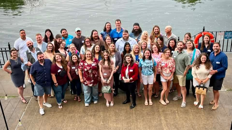 Large group posing for a photo on a lakeside dock, with water in the background and one person highlighted in red.