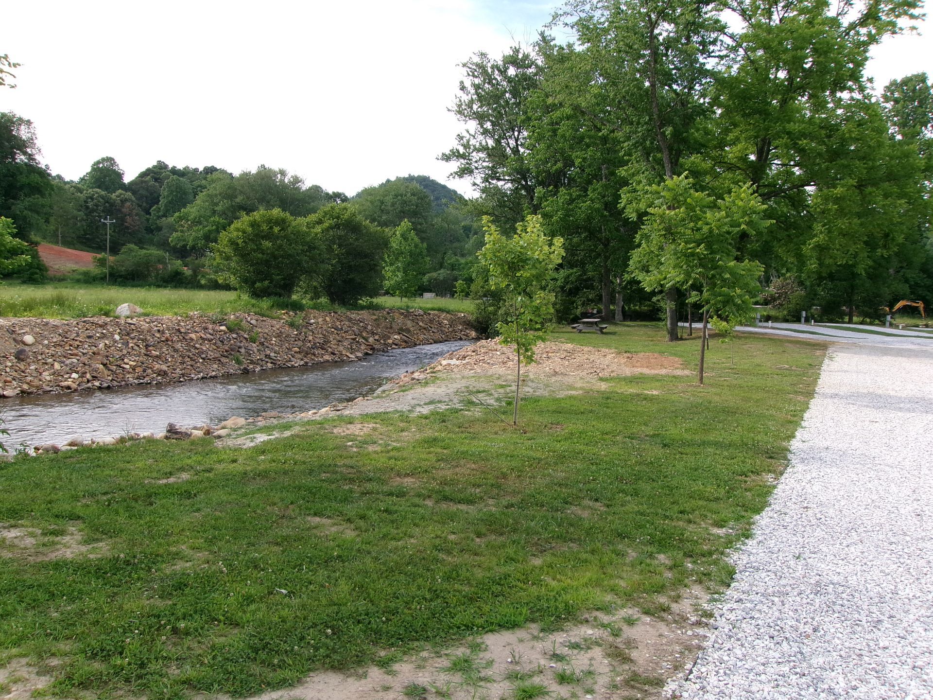 Grassy area with a gravel path, a dry creek bed lined with stacked logs, and trees under a cloudy sky.