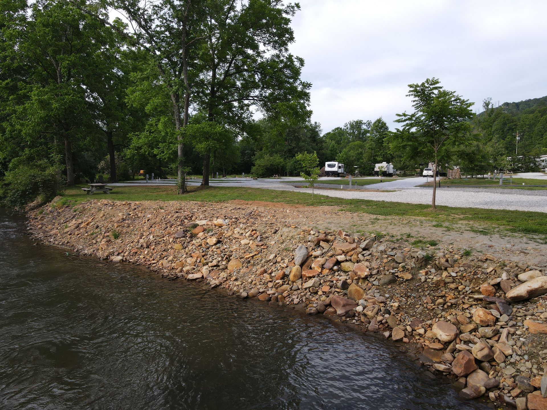 A campsite alongside a river. Brown rocks line the riverbank, while RVs are parked among trees.