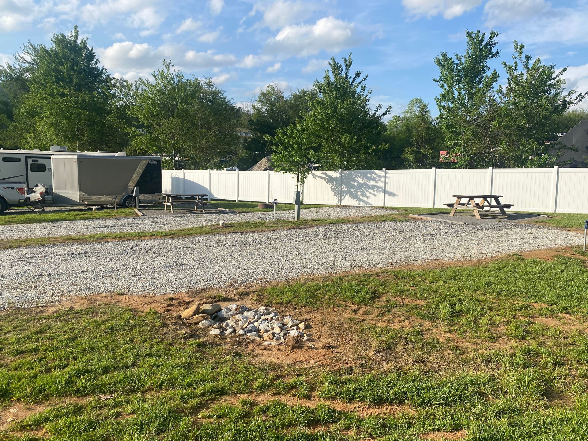 Campsite with gravel RV parking, picnic tables, a fire pit, and a white fence under a blue sky.