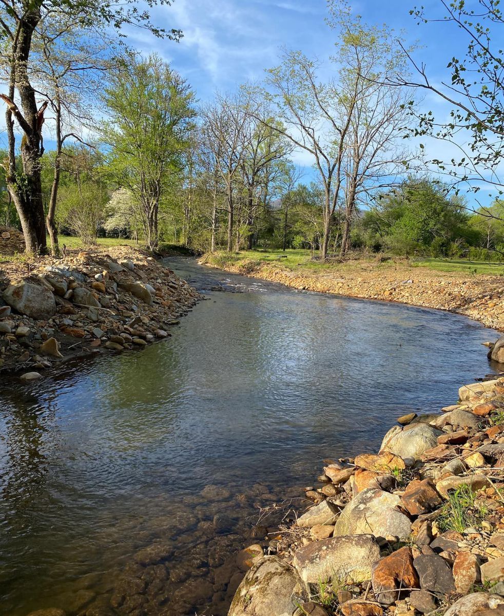 A serene river flows through a sunny landscape, framed by rocky banks and lined with trees under a partly cloudy sky.