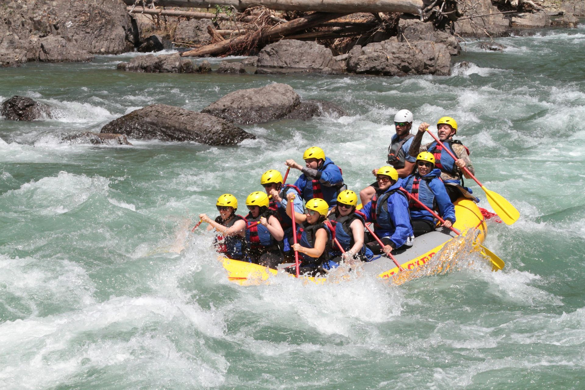A group of people in a yellow raft navigate a whitewater river, paddling through rapids. They wear helmets and life jackets.