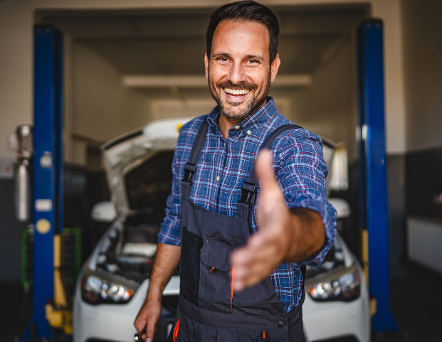 Smiling mechanic in blue shirt and overalls reaching out in a garage with a car on a lift behind him | Kars Performance
