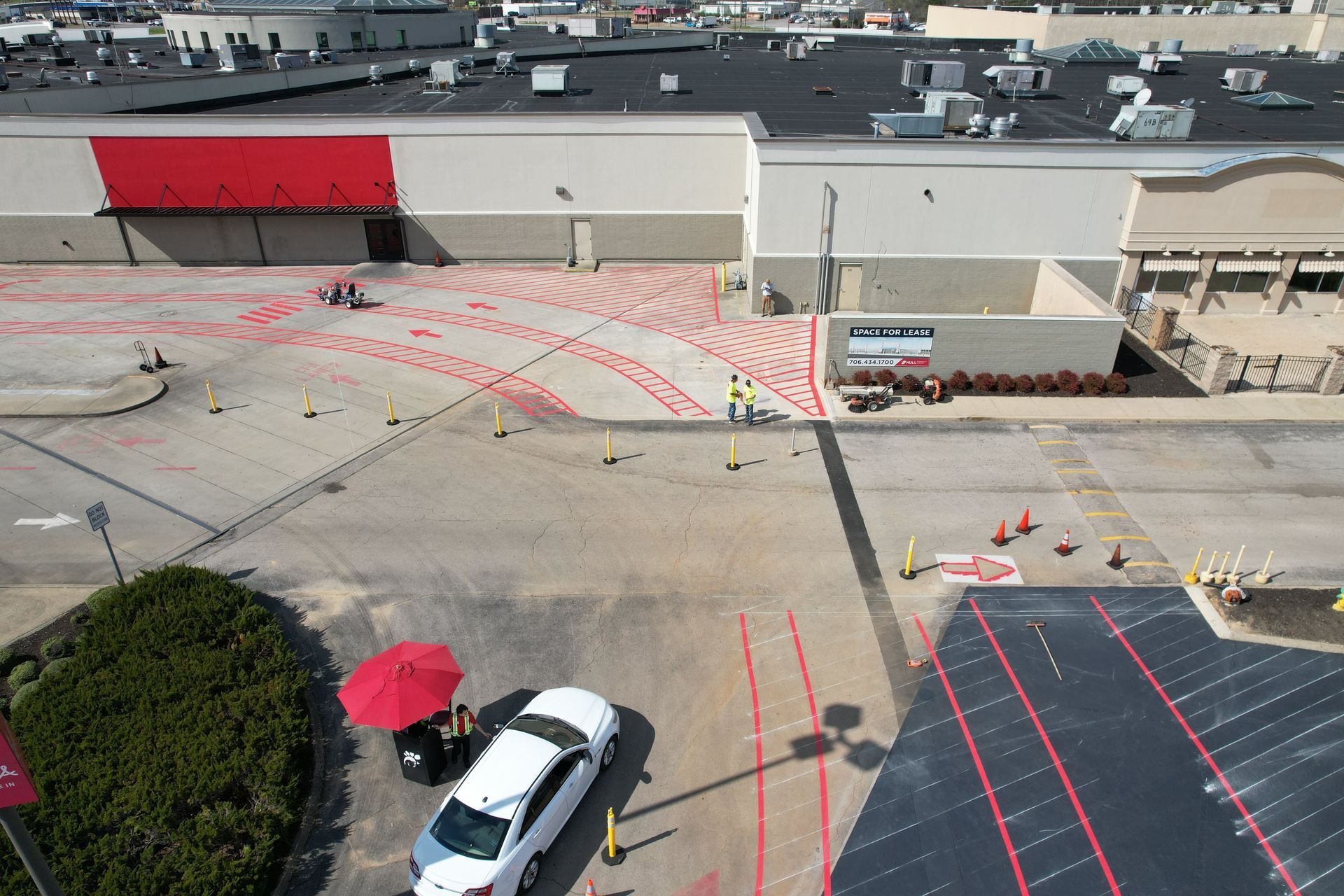 Aerial view of an industrial loading area with red-striped pavement, trucks, and a red umbrella near a white car