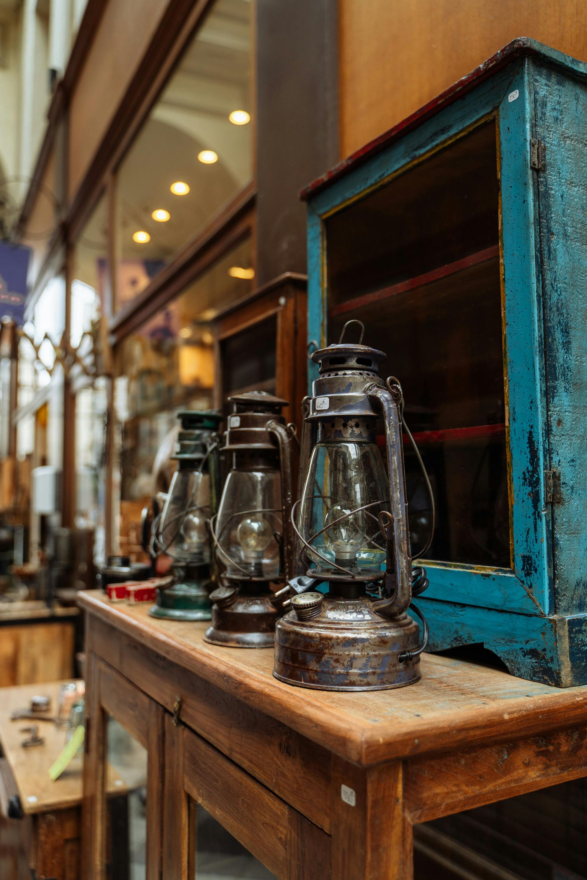 Vintage lanterns on a wooden table beside a teal cabinet in a warm shop interior